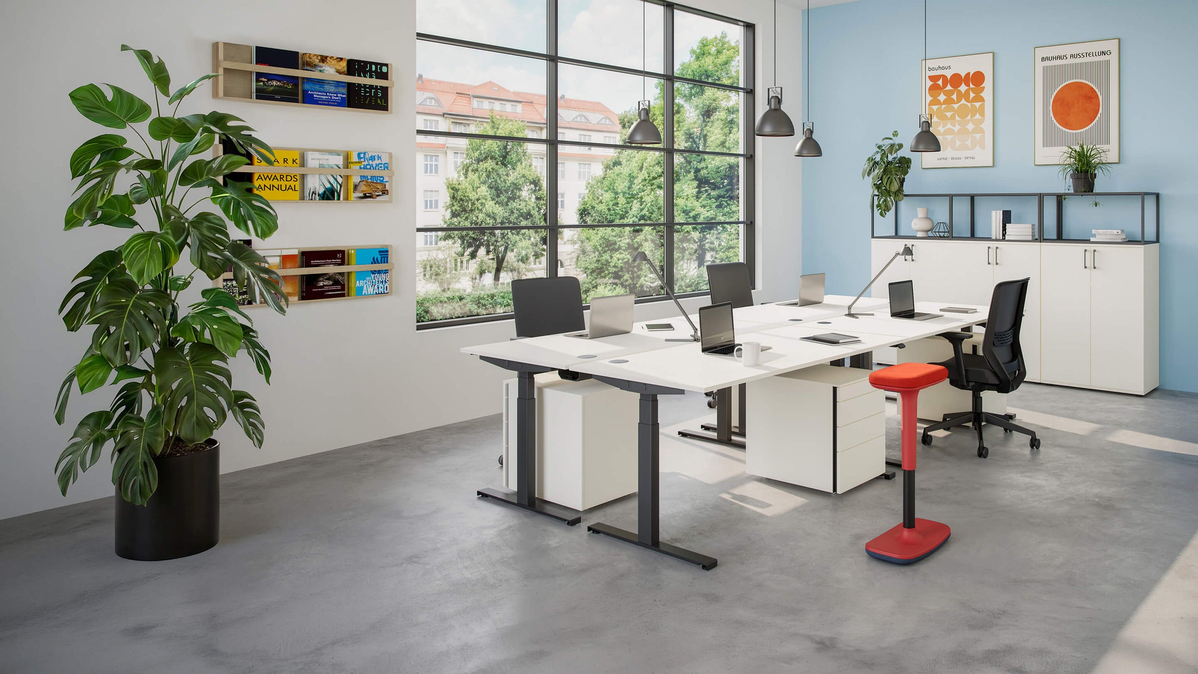 Modernes, helles Büro eingerichtet mit hali Möbel- zu sehen ist ein großes Fenster, weißen Schreibtischen, ergonomischen Bürostühlen und Stauraummöbeln. Pflanzen, Wandregale, Poster und ein roter Stehhocker setzen wohnliche Akzente.